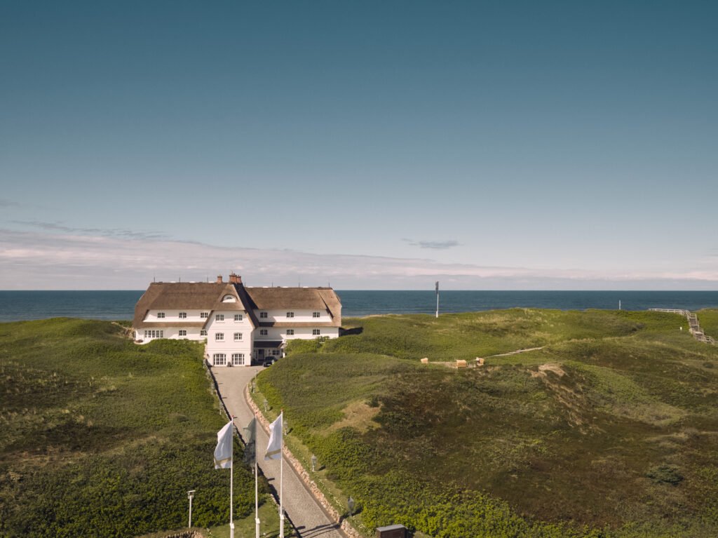 Söl’ring Hof: weißes Strohdachhaus, Dünen, Meer, blauer Himmel.