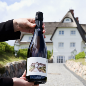 Eine Person hält eine Flasche „Terroirs d’Ecueil“ in der Hand; im Hintergrund sind das Haus und die grüne Umgebung zu sehen.
