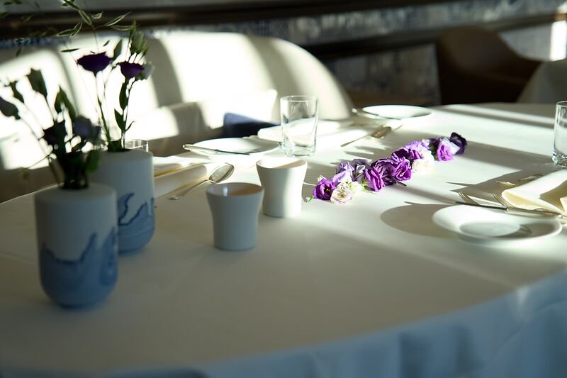 Runder Tisch mit weißem Tuch, lilafarbenen Blumen und eleganter Tischdekoration im Sonnenlicht.