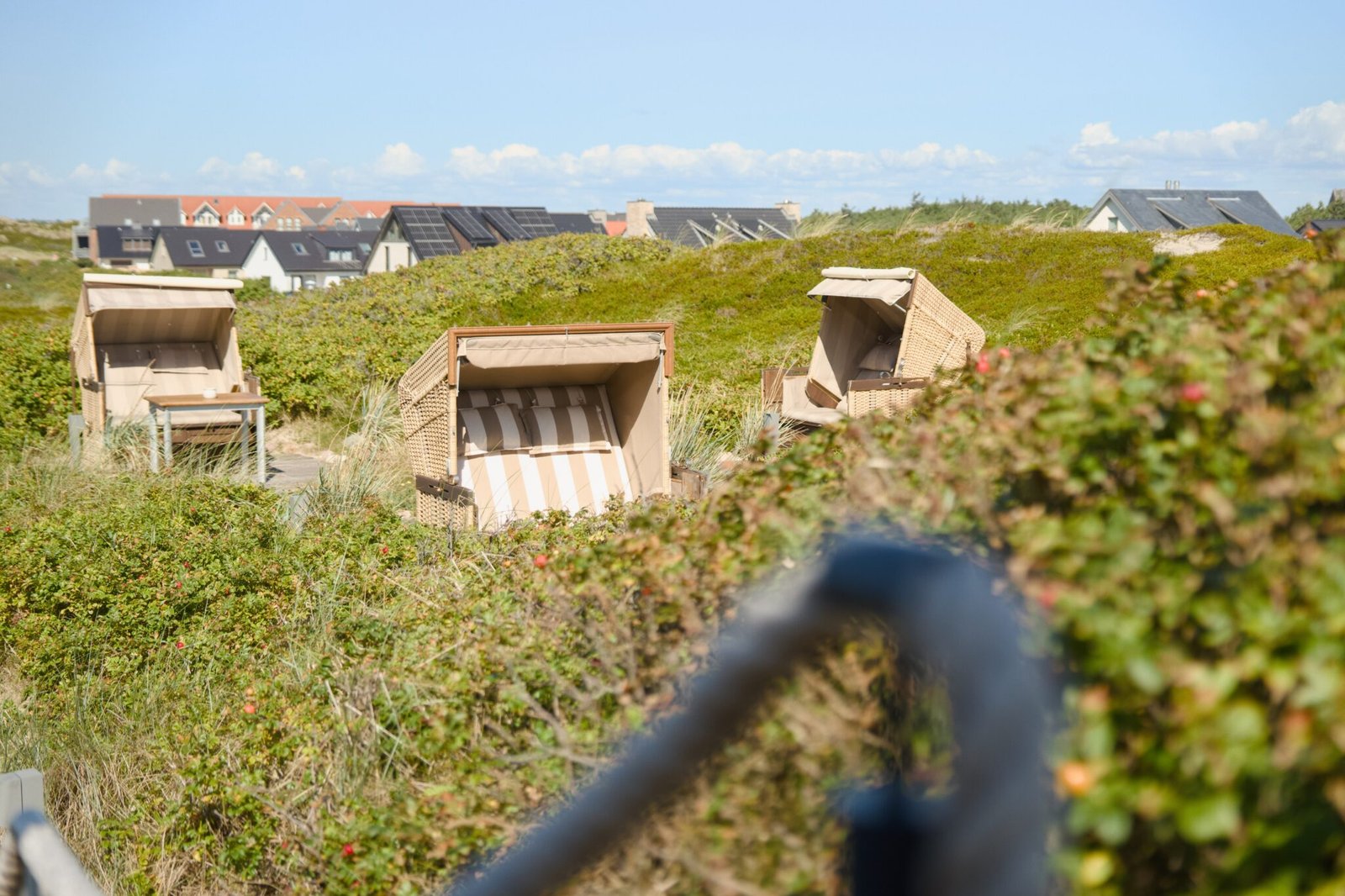 Drei gestreifte Korbstühle inmitten grüner Dünen; dahinter Häuser und Sonnenkollektoren.