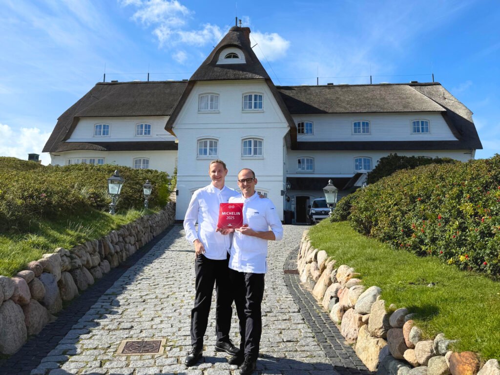 Zwei Köche halten eine rote Michelin-Plakette vor einem reetgedeckten Gebäude.