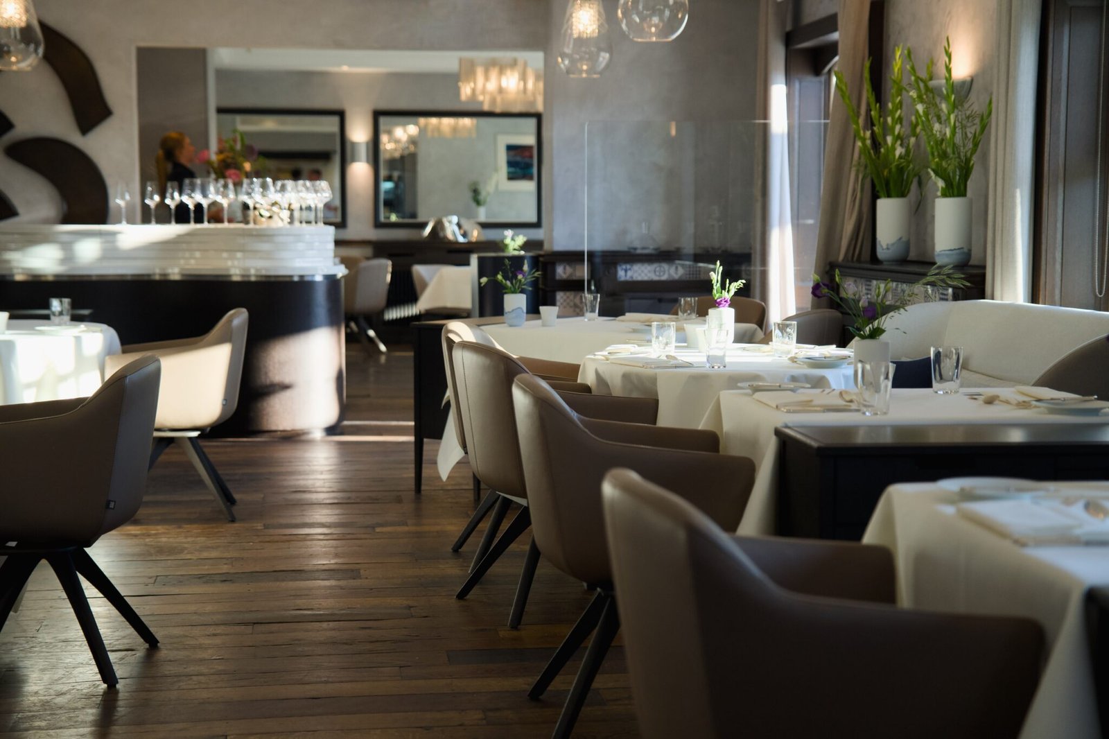 Modernes Restaurant: weiße Tische, beige Stühle, Holzböden, Pflanzen, Sonnenlicht.