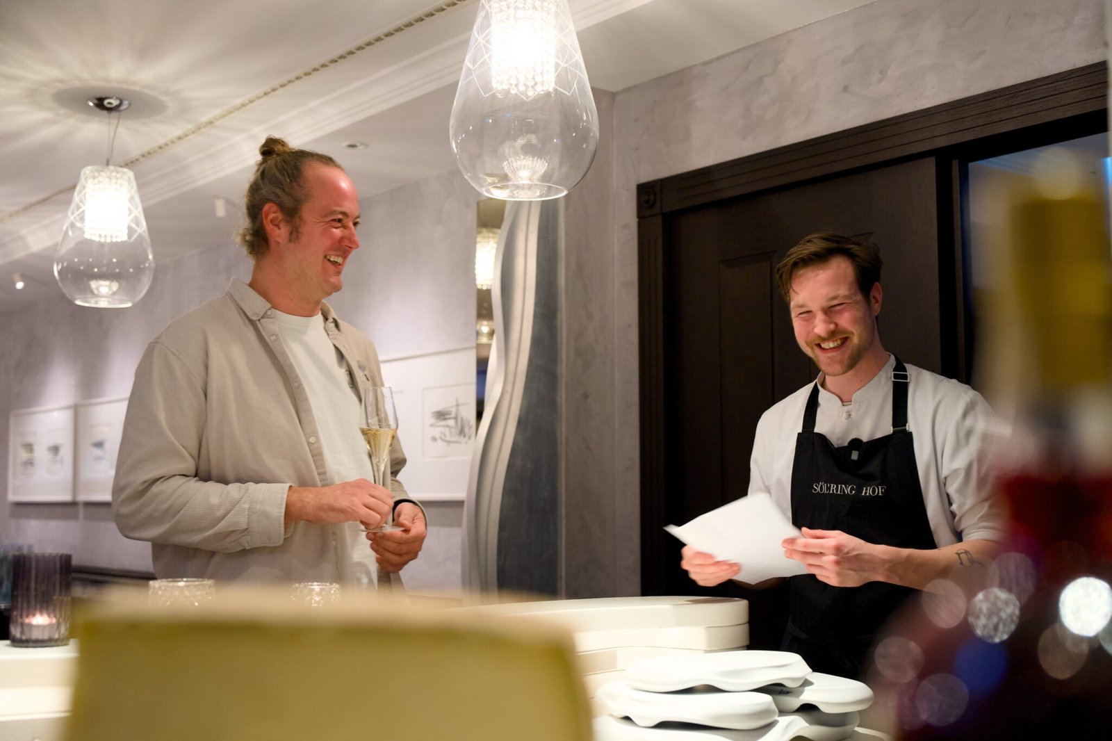 Two men smile and talk in a modern restaurant, stacked plates nearby.