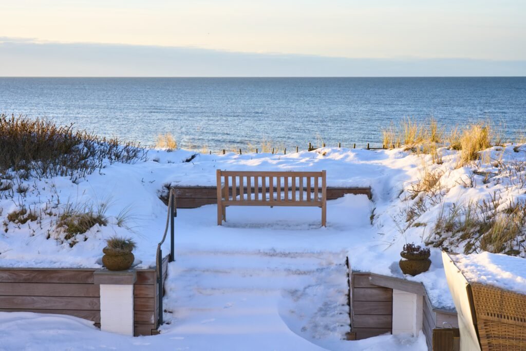 Holzbank auf verschneiter Terrasse, ruhiges Meer vor sich, klarer Himmel.