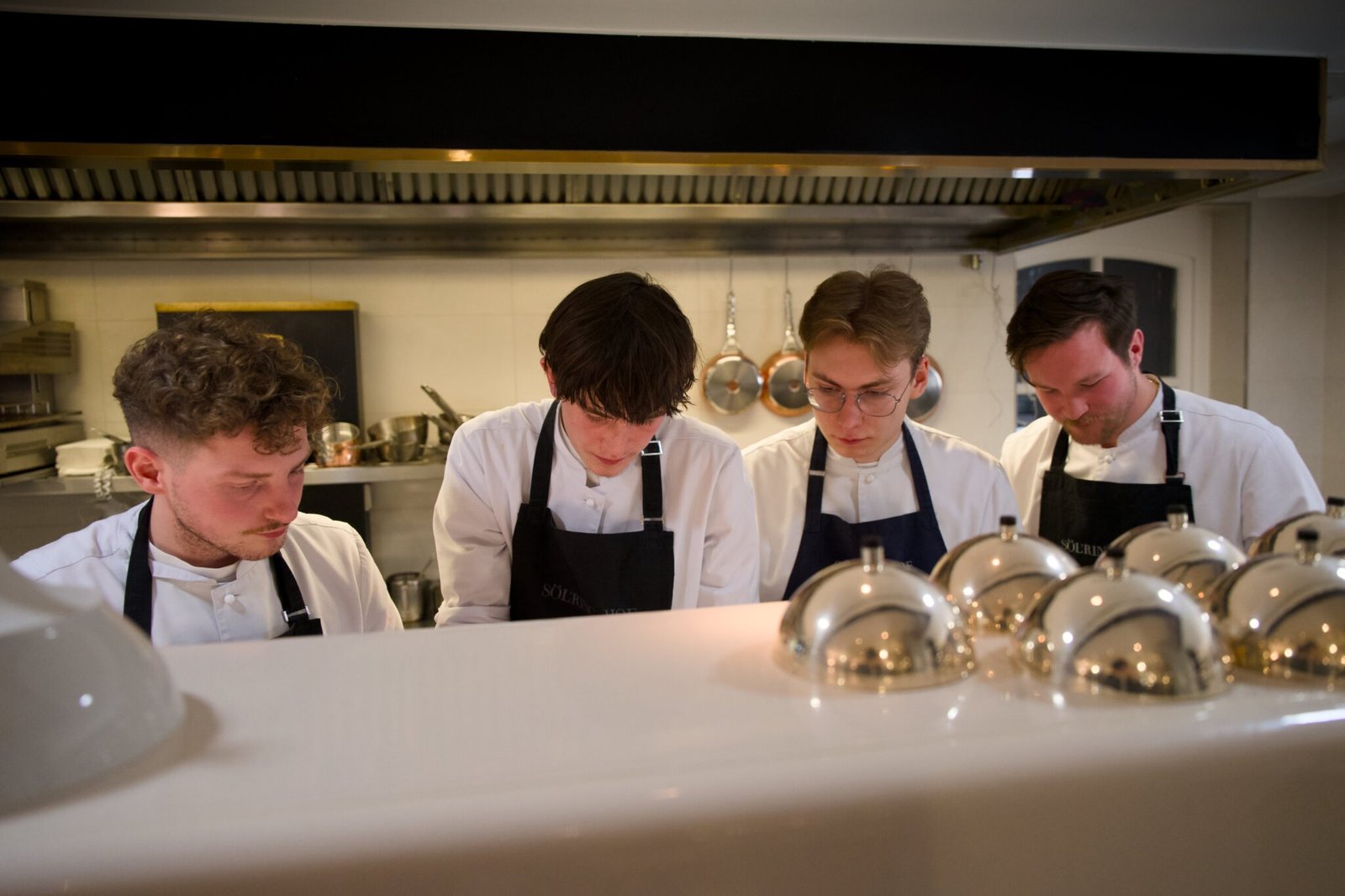 Four chefs cooking together; pans hang, silver domes sit nearby.