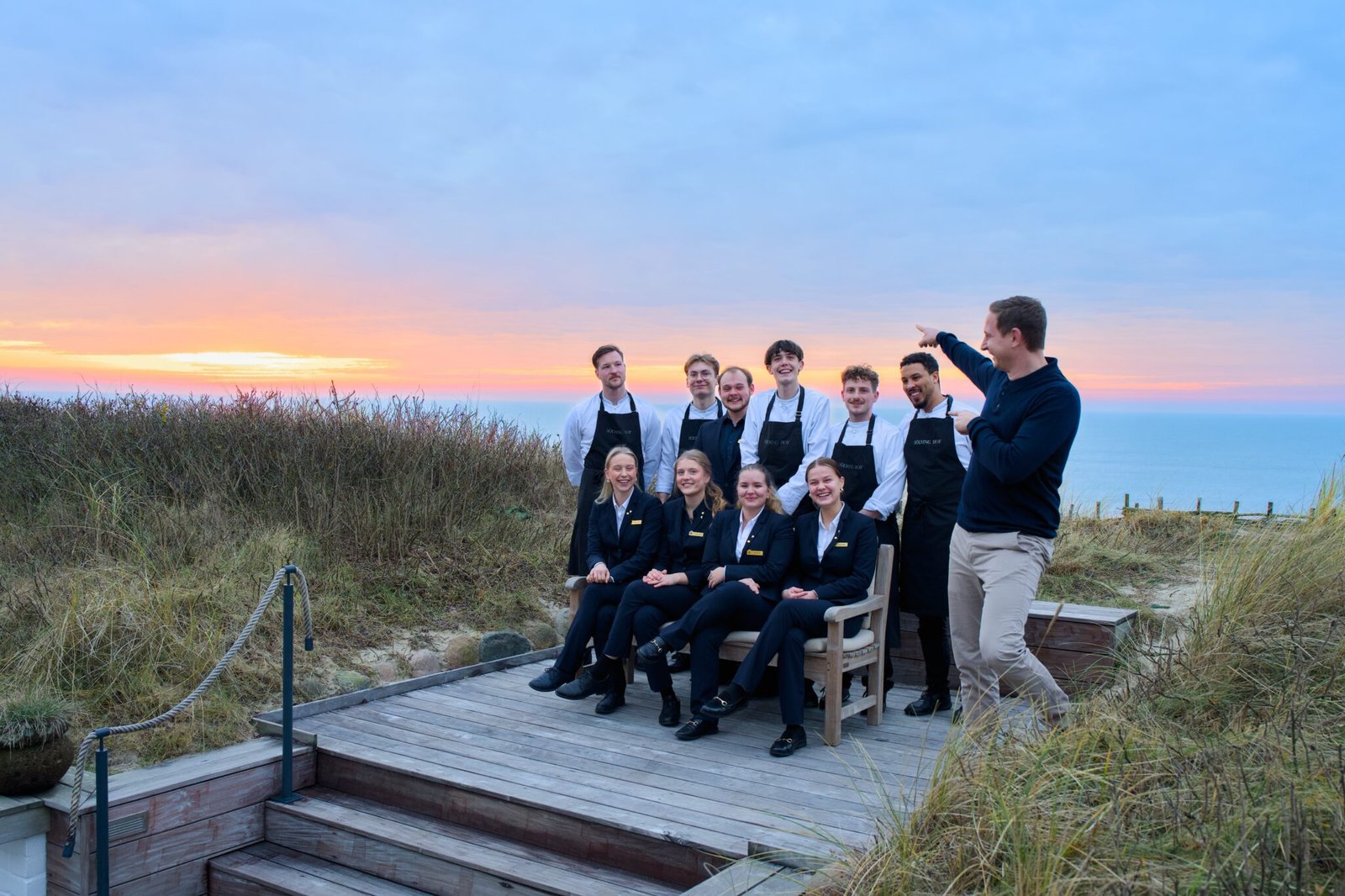 Ten people on steps by ocean at sunset; one points.