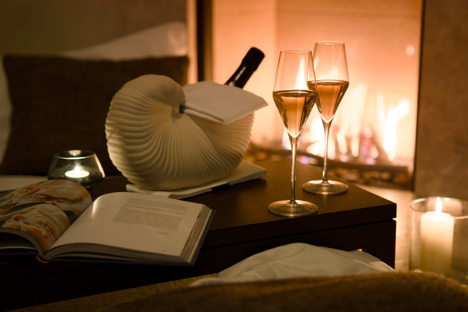Champagne, wine, book, and candles on table before fireplace—cozy.