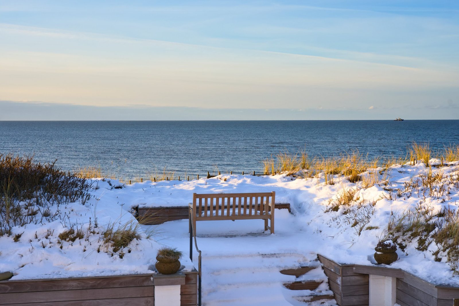 Bench faces snowy dunes, ocean, blue sky, distant ship visible.