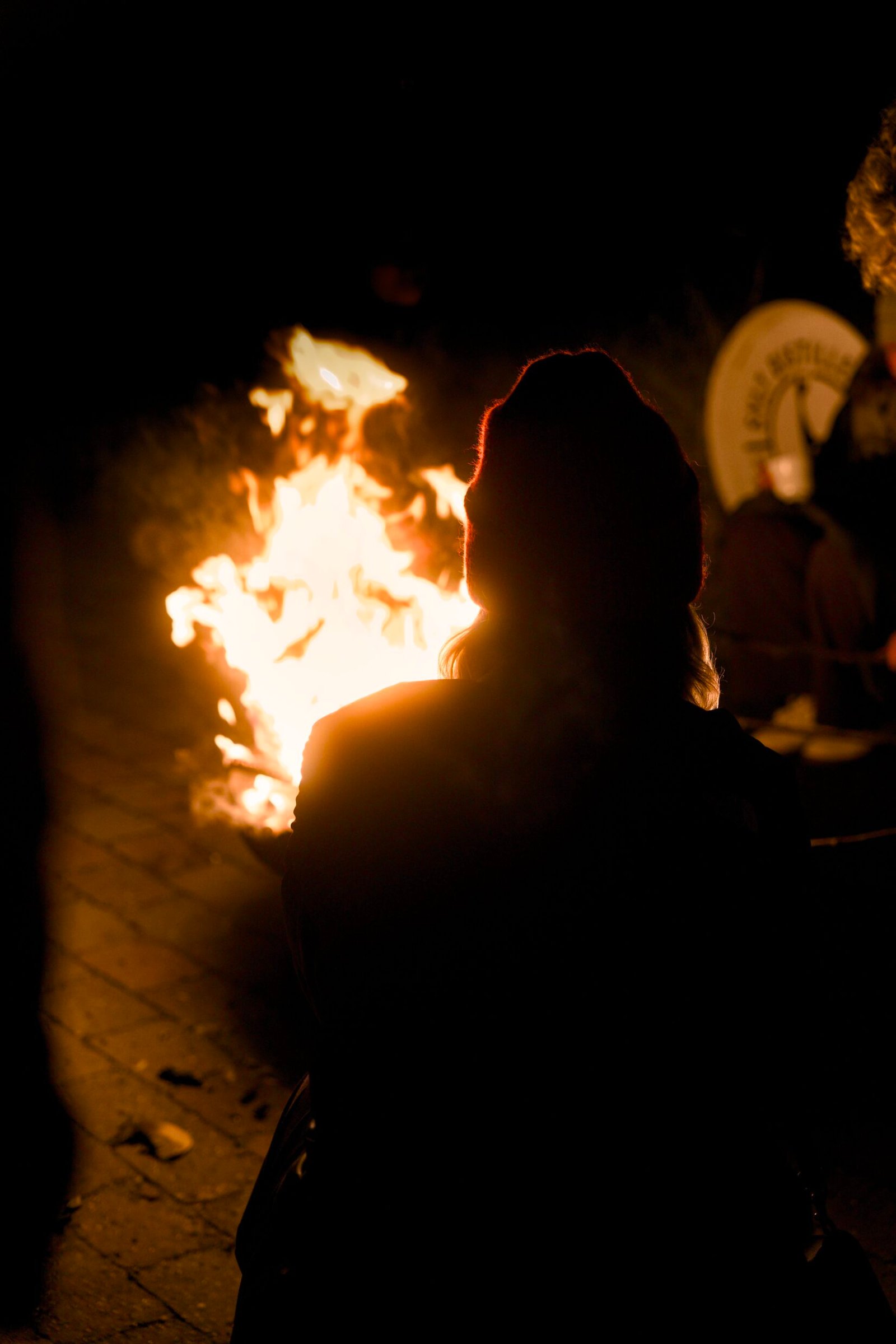 Beanie-wearer sits by night fire pit, warm glow.