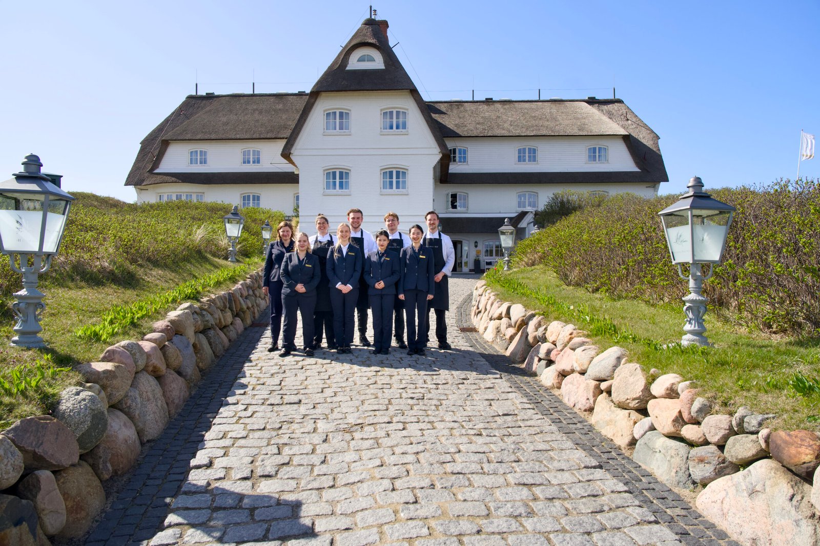 Acht Personen in formeller Kleidung stehen auf dem Kopfsteinpflaster vor einem strohgedeckten Gebäude.