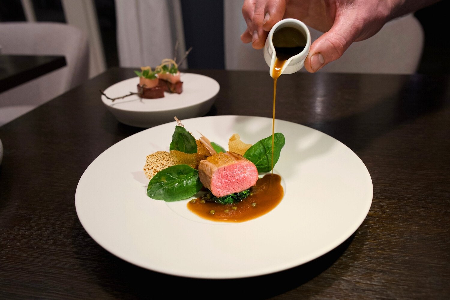 Eine Hand gießt Soße über Steak und Gemüse; dahinter ein verschwommener Teller.
