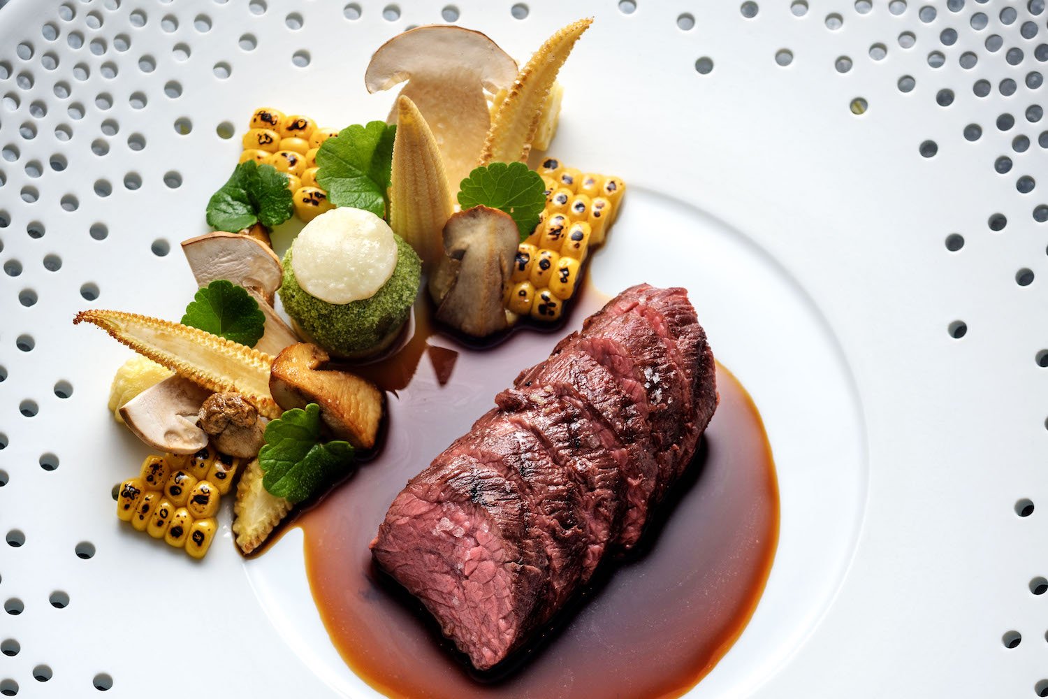 Seared steak with brown sauce, corn, mushrooms, and greens plated.