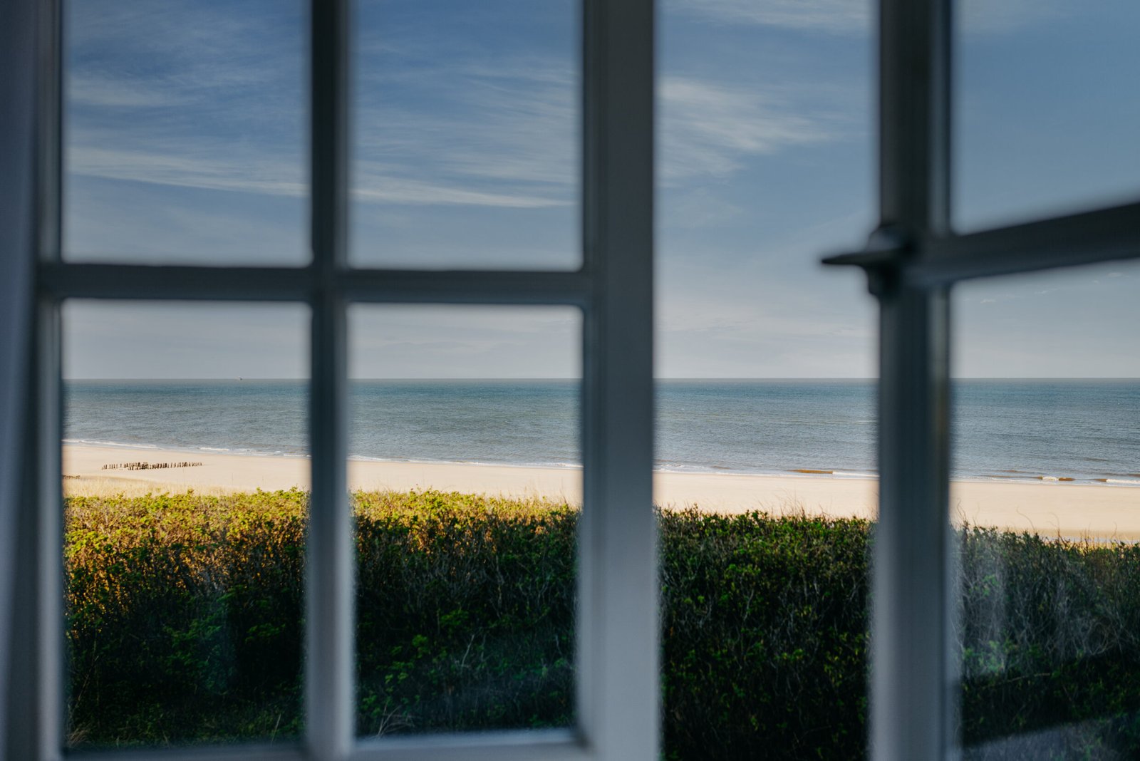 Window frame in focus, beach, greenery, and calm sea beyond.