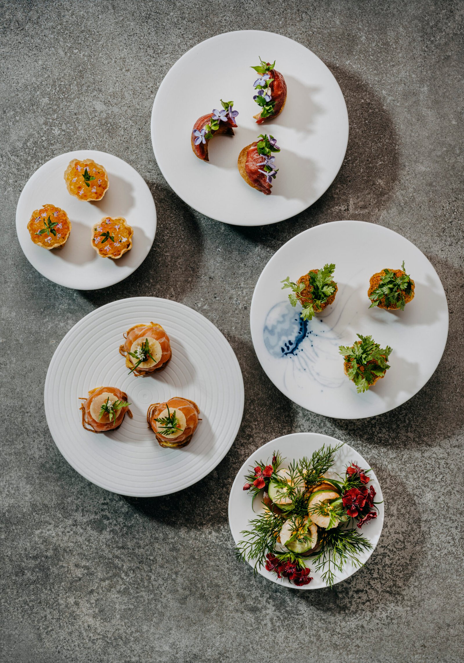 Five white plates with assorted garnished appetizers on a gray surface.