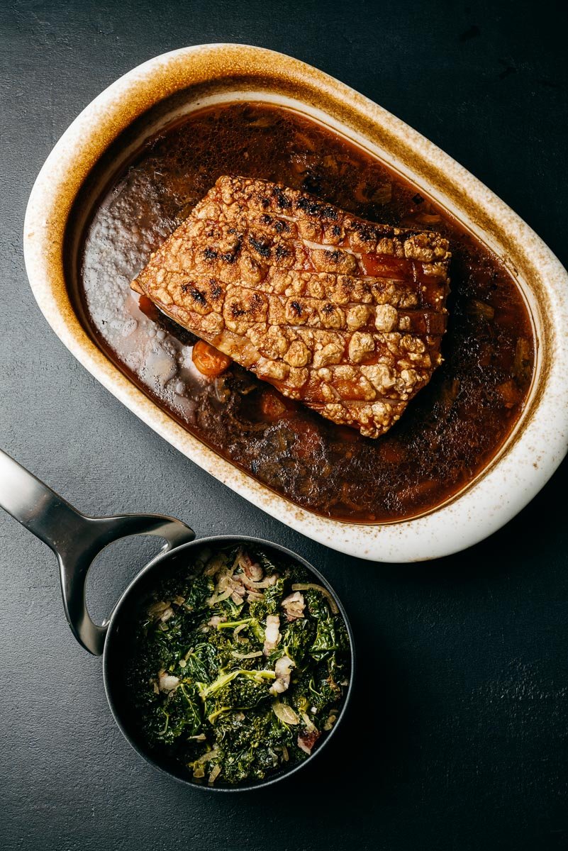 Roast pork belly with crackling, greens, and bacon on dark surface.