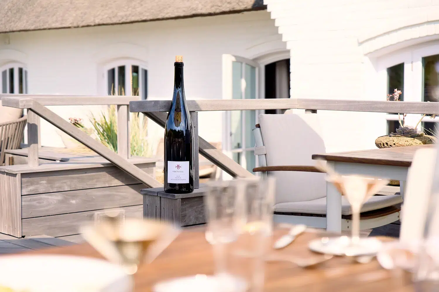 Weinflasche auf einem Terrassenpfosten, Stühle und ein weißes Gebäude im Hintergrund.