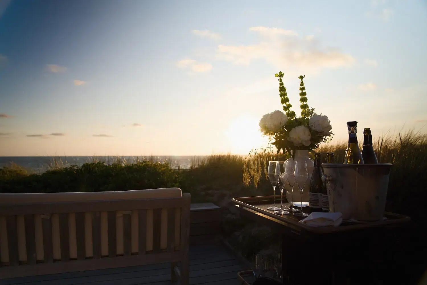 Bench, table with drinks, flowers; sunset over ocean and grass.
