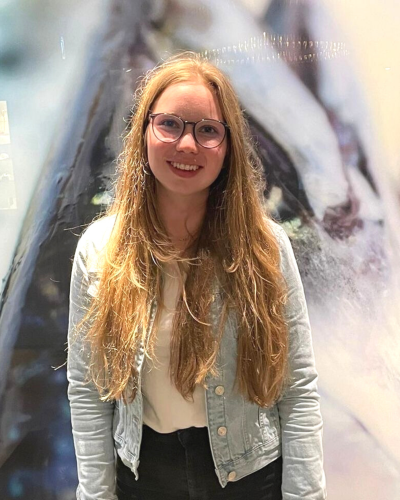 Lächelnde Frau mit Brille, Jeansjacke, unscharfer Hintergrund.