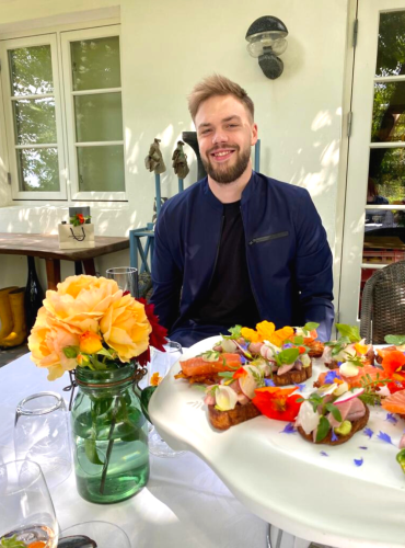 Blonder Mann mit Bart speist im Freien, Blumen und Essen vorhanden.