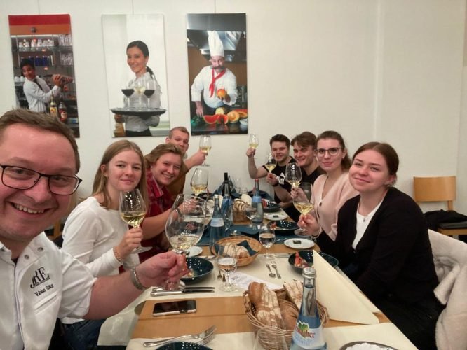 Neun Personen stoßen lächelnd mit Wein an einem Restauranttisch an.